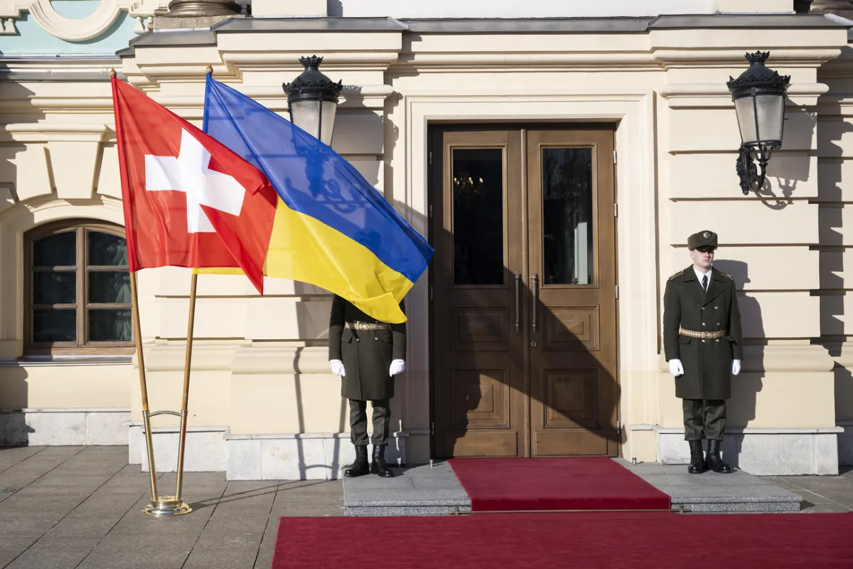 Two soldiers are guarding a building, with the flags of Ukraine and Switzerland in front of them